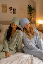 Two people sit together on a bed wearing Sunlit Meadow and Blushing Sky Sleep Hoods.