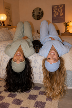 Two people lie side by side on a bed wearing sage green and blue Sleep Hoods, with a small dog between them.